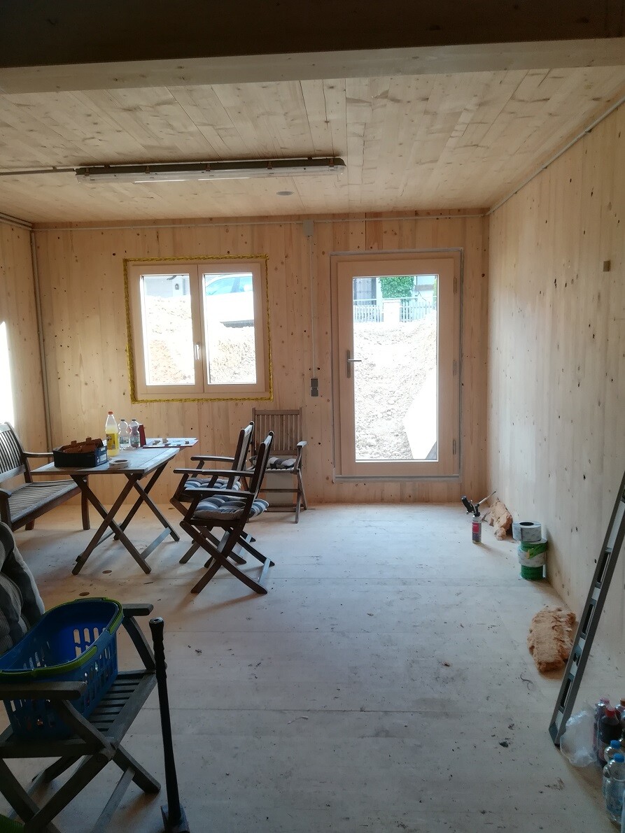 Shell structure made of wood with dining table, chairs, construction tools, window and door leading outside.