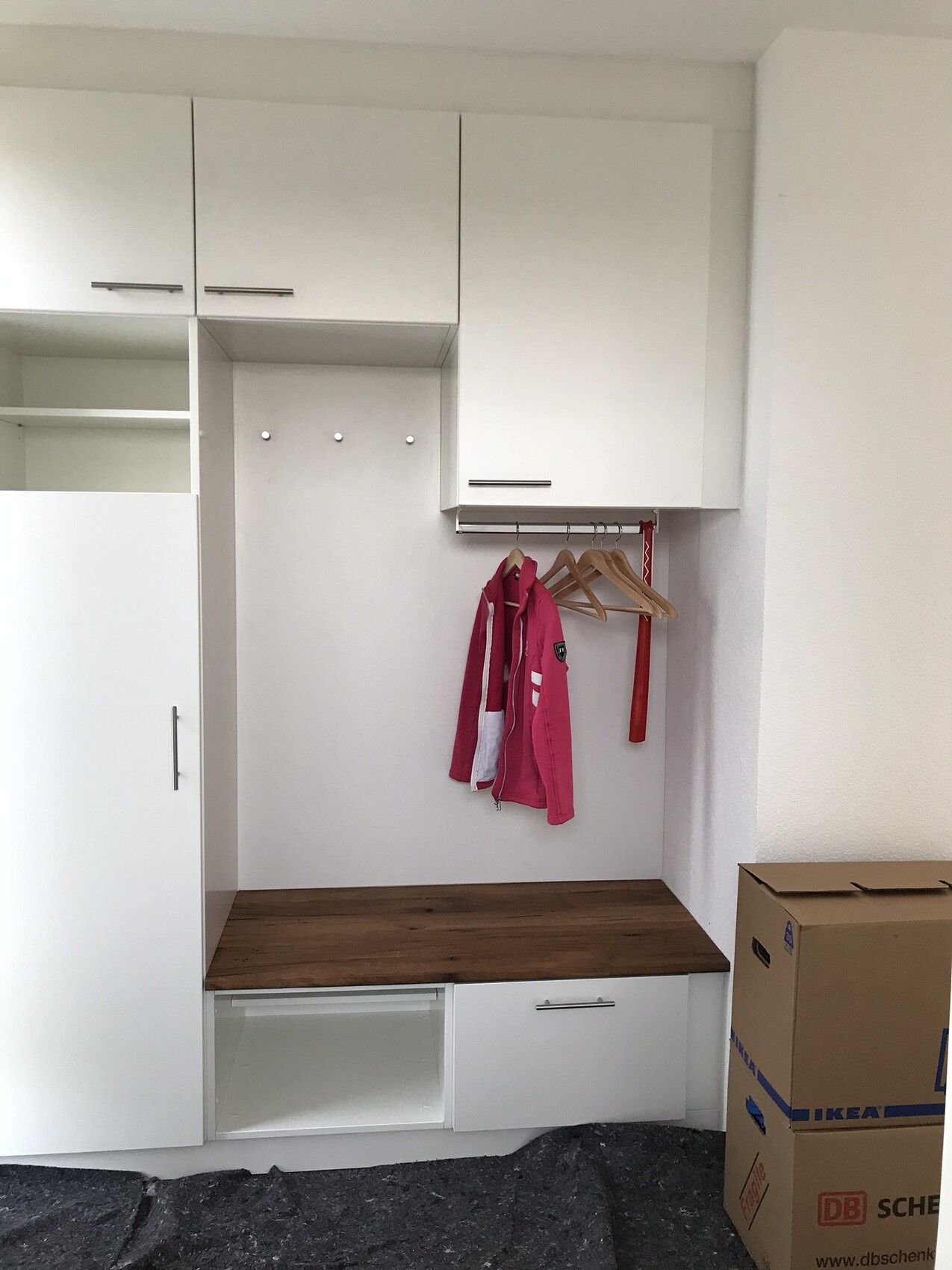 White built-in wardrobe with wooden shelf, hook rail and pink jacket; moving boxes on the right.