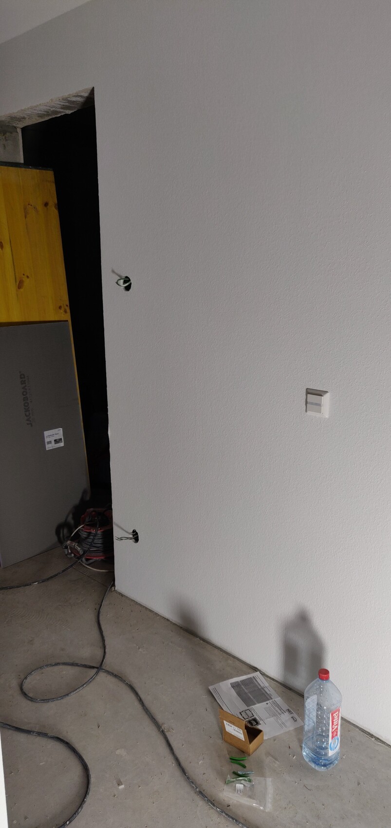 White interior wall with hole for electrical outlet, loose wires, construction equipment, cardboard box, and water bottle on the floor.