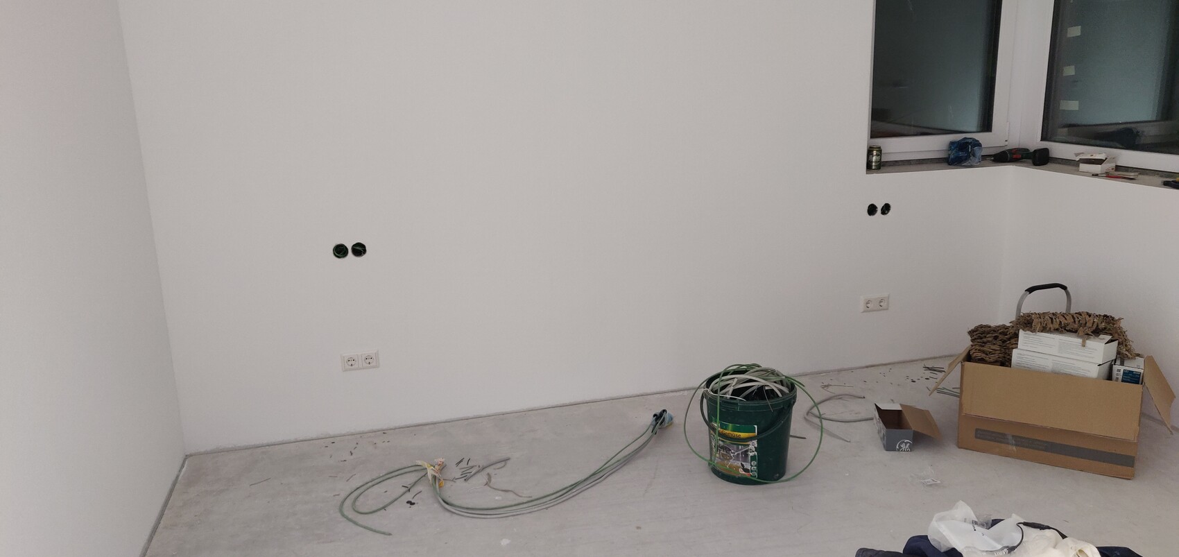 Renovation room with white walls, open electrical outlets, cables, boxes, and tools on the windowsill.