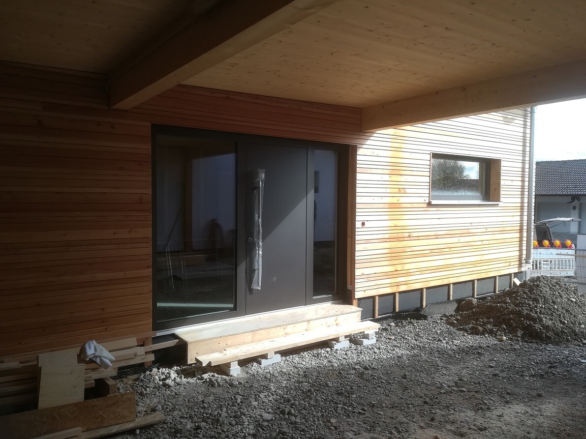 Wooden house under construction: dark door front, horizontal wood cladding, construction site with debris.