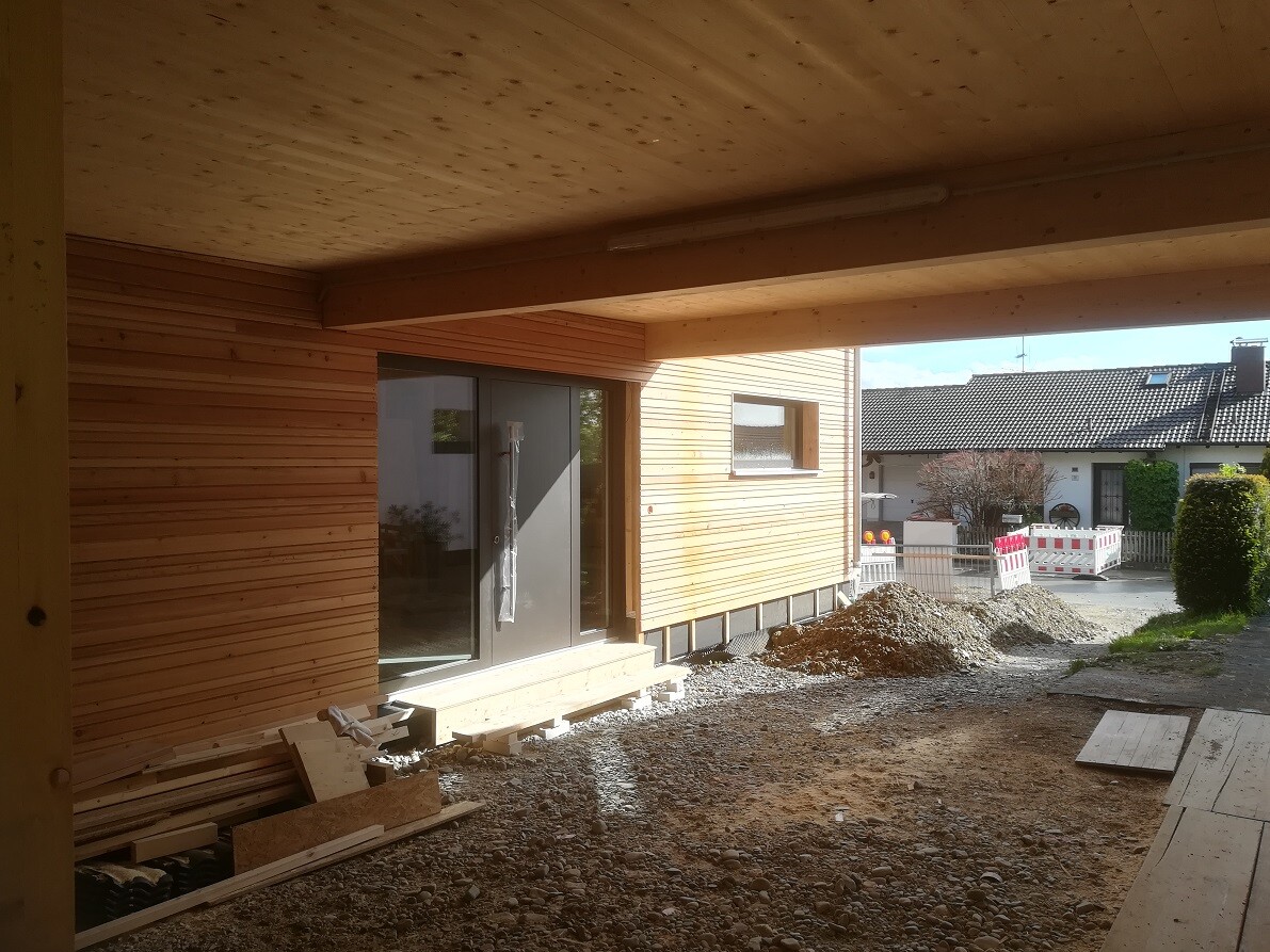 Wooden house under construction with glass door, wood cladding, gravel floor and construction equipment in the foreground.