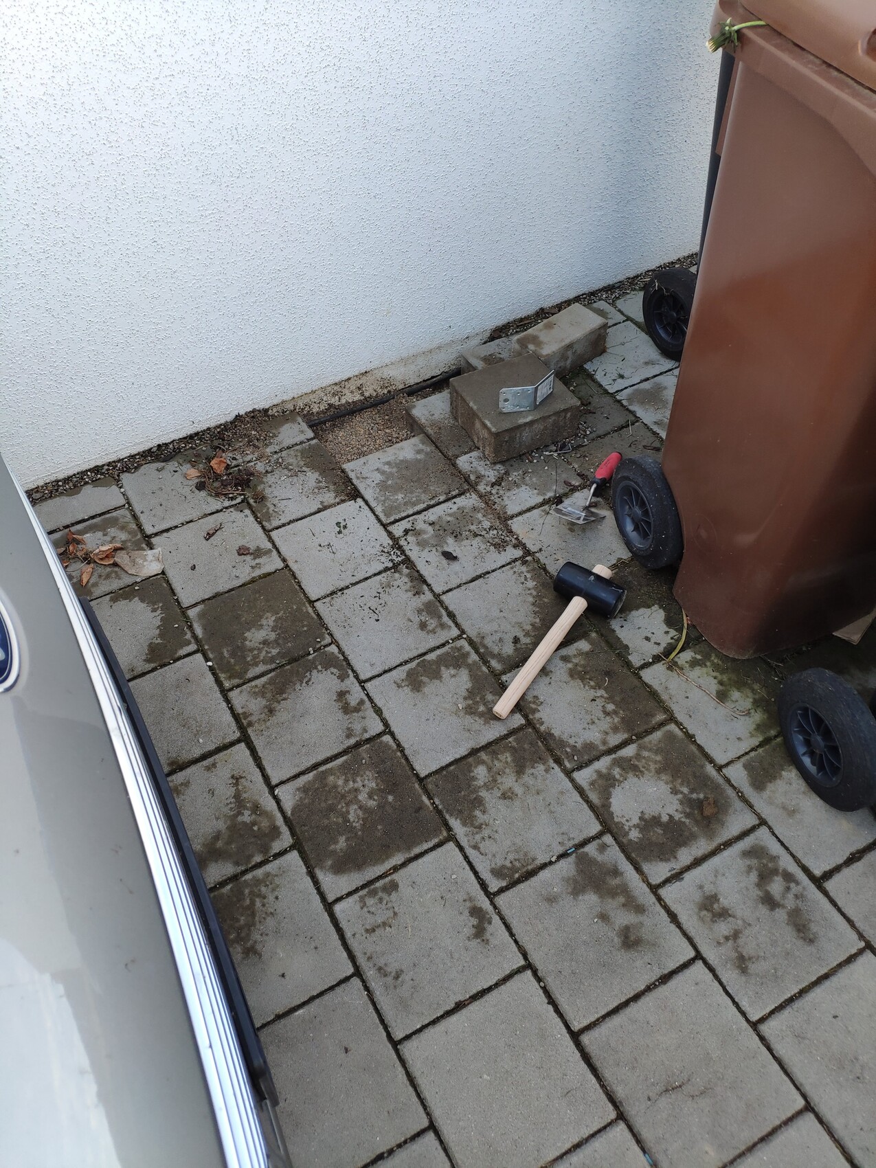 Paved patio next to white wall; brown trash container, stones and tools scattered around.