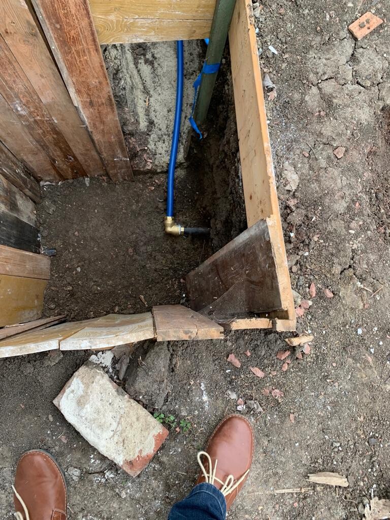 Open construction pit with blue PEX water pipe and wooden formwork; feet in the foreground.