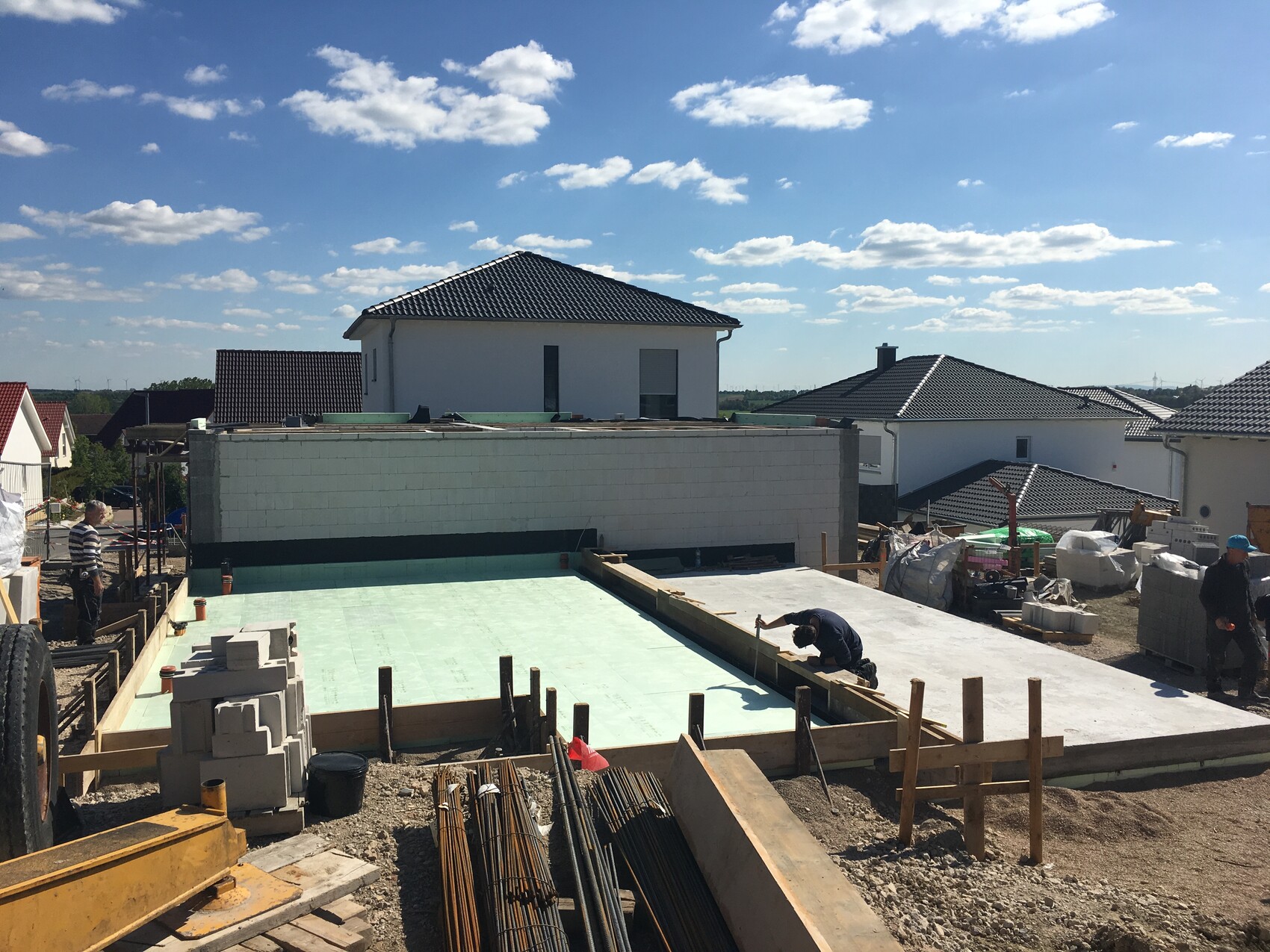 Unfinished construction site with green concrete floor, stones, wooden planks, and workers in front of houses.