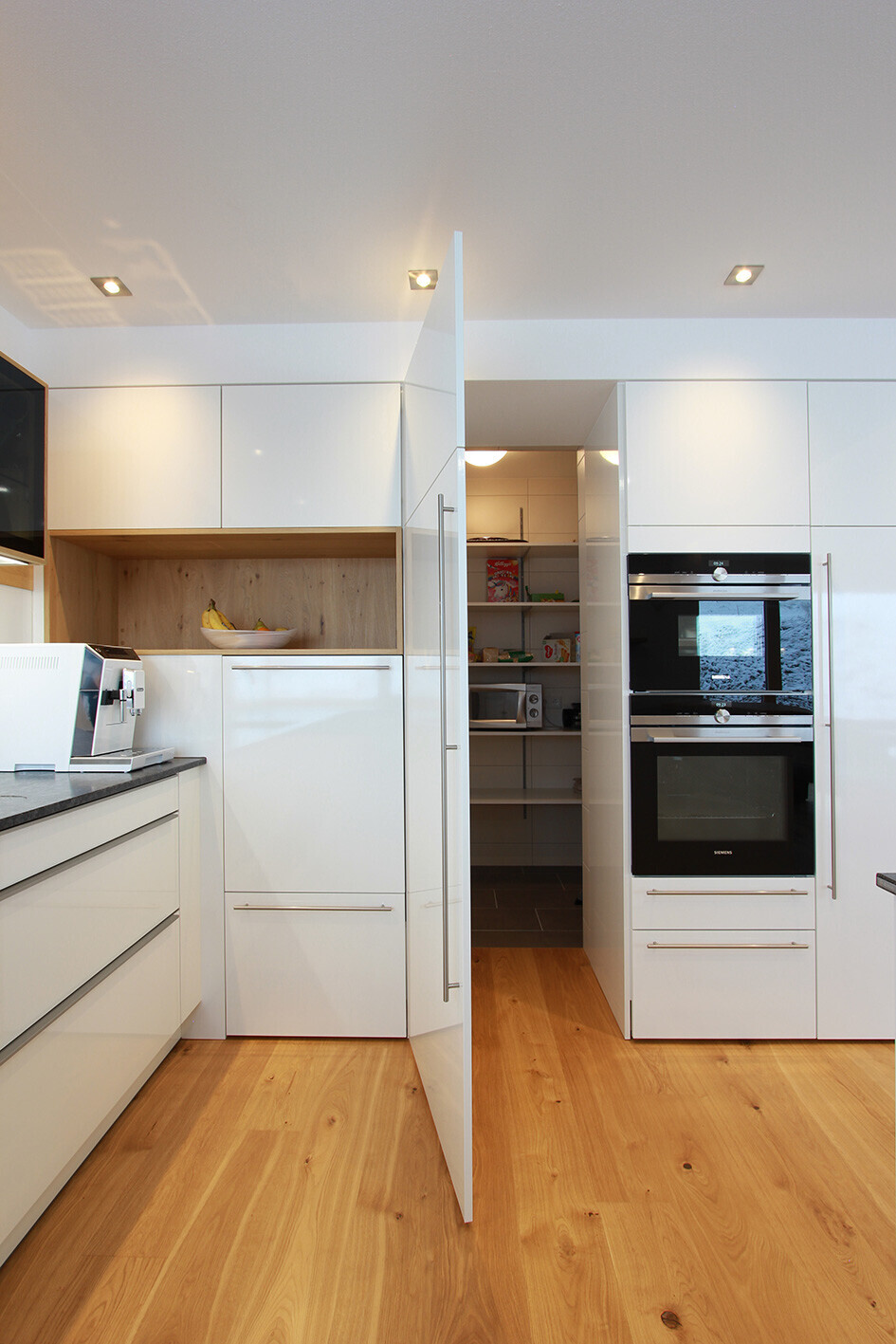 Modern bright kitchen with open pantry, integrated ovens, and wood flooring.