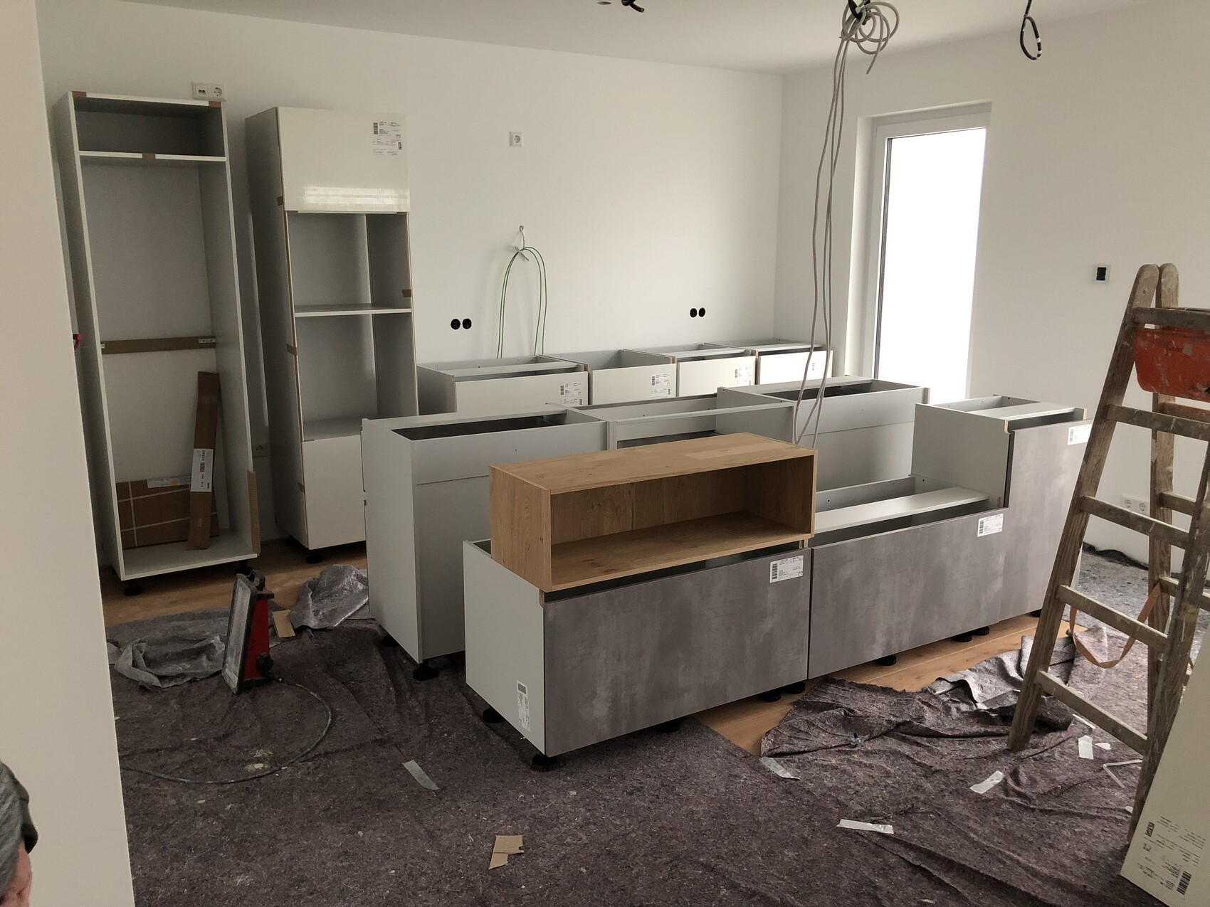 Kitchen under construction: grey base cabinets, open cabinets, ladder, hanging cables from the ceiling.