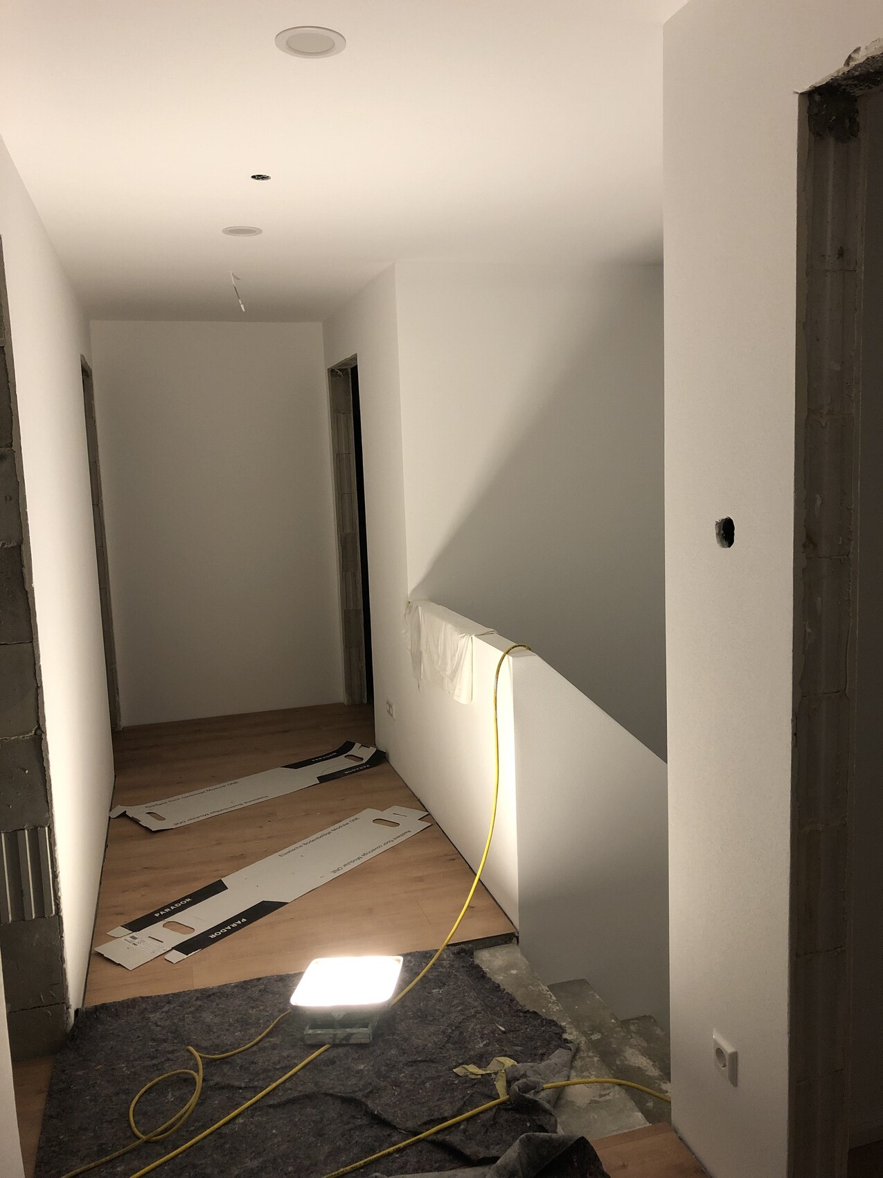 Bright hallway under construction: white walls, loose panels, yellow cable and work light on the floor.