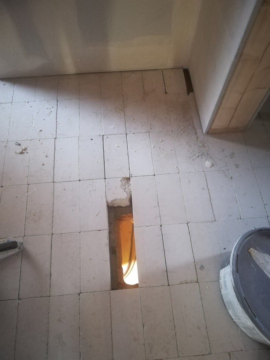 Partly removed floor tiles; long rectangular hole shows orange light and pipes underneath.