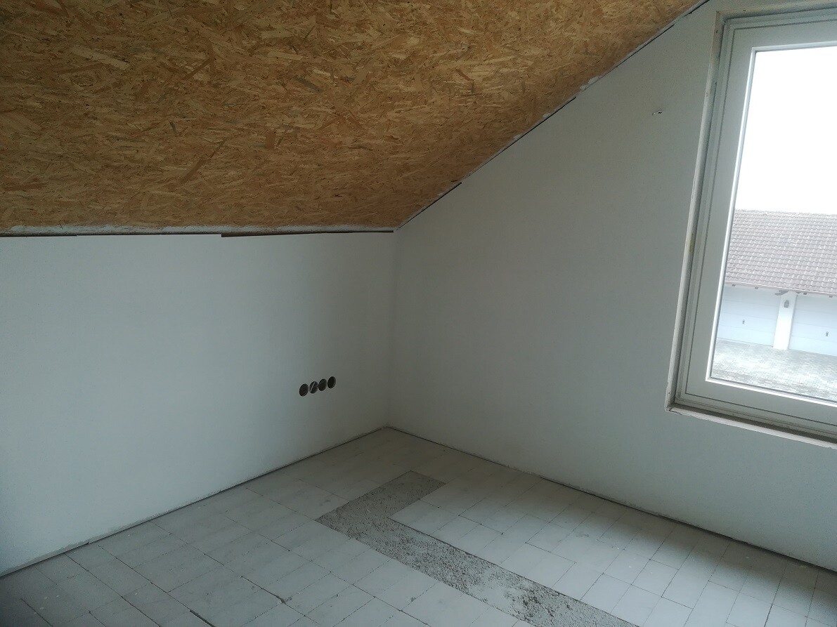 Empty space in attic with sloped OSB ceiling, window on the right, white walls, five round electrical outlets.