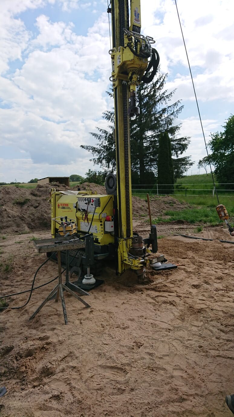 Gelbe Bohranlage/Bohrgerät steht auf einer Baustelle mit Sandboden; Hintergrund Felder und Bäume.