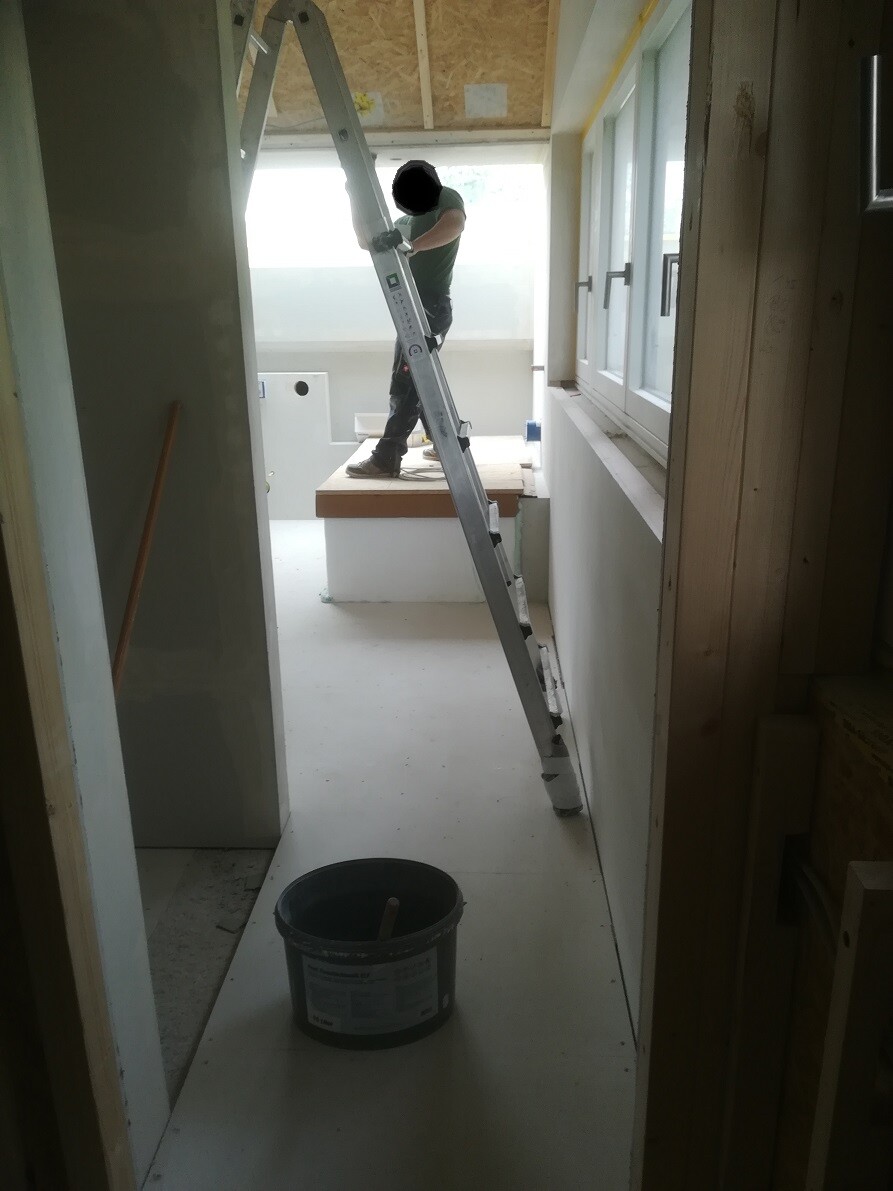 Man standing on a ladder inside a construction room working on the ceiling; bucket on the floor