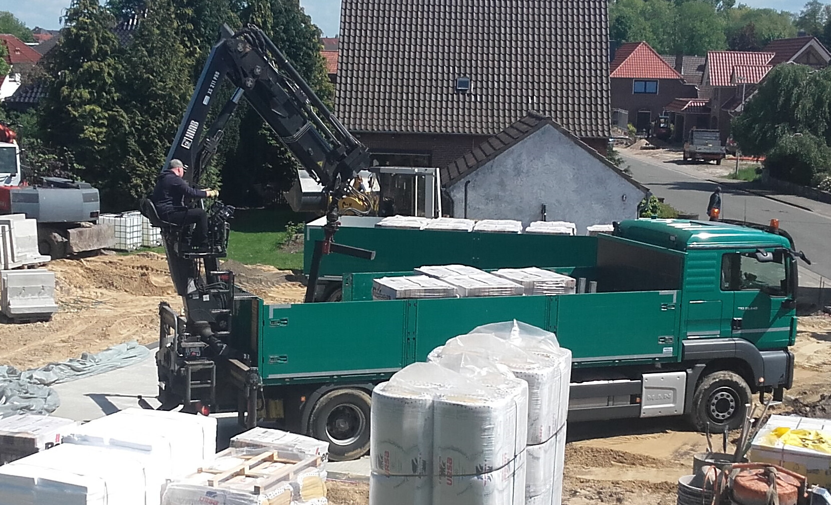 Green truck with crane at construction site, worker operating crane arm; stacked building materials.