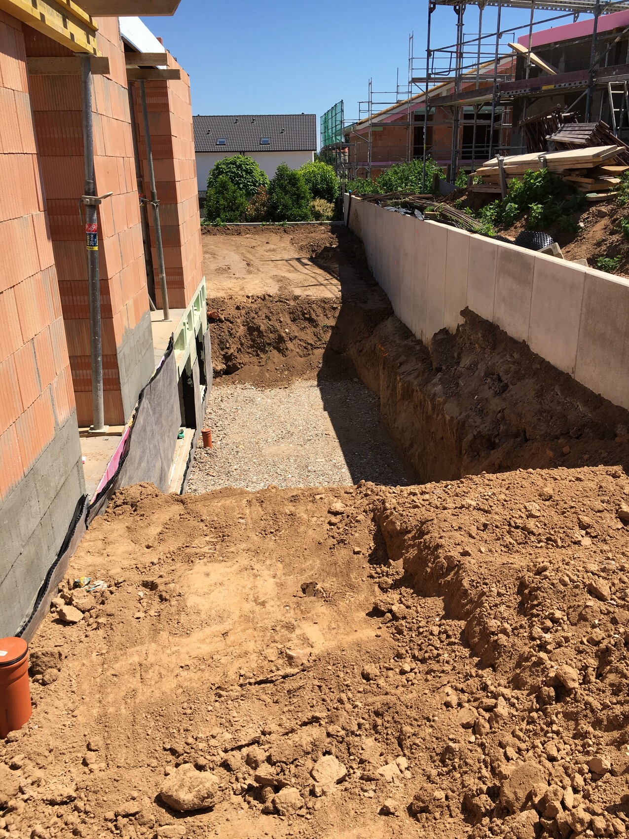 Excavation next to unfinished brick house on the left, concrete wall on the right, scaffolding and wood stack in the background.