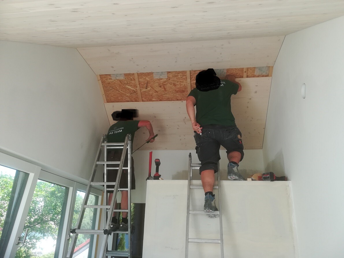Two construction workers on ladders attaching wooden panels to a sloped ceiling inside.