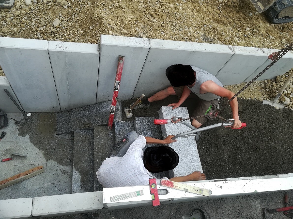 Two construction workers installing concrete steps on a staircase, with crane hooks and tools.