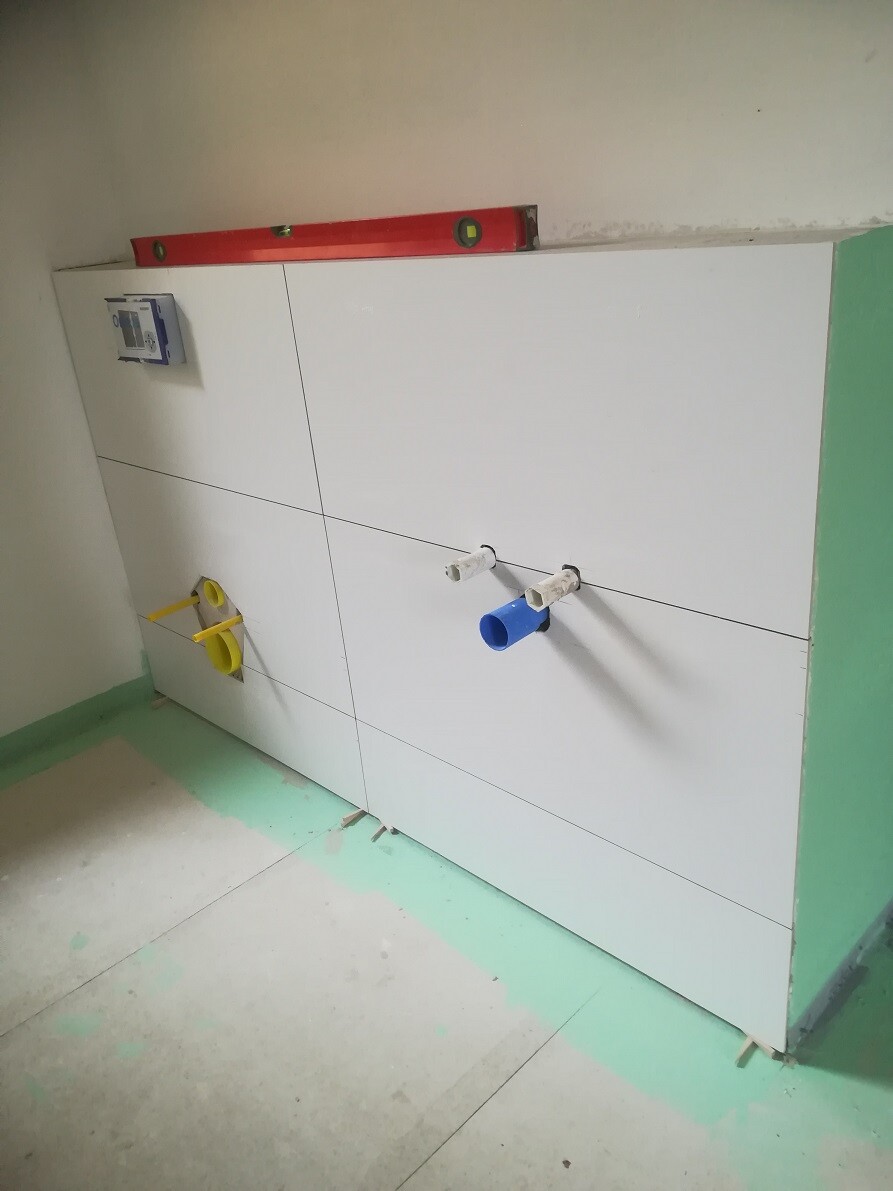 White tiled wall under construction with blue water connection and yellow electrical outlets; level placed on top.