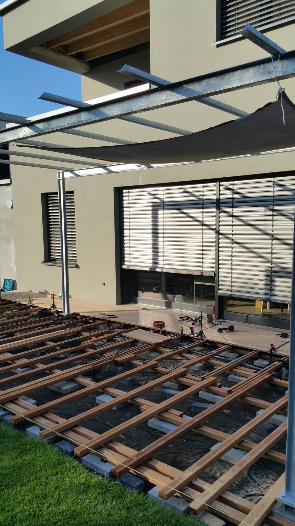 Wood terrace substructure in front of modern house; metal frame with sun sail, tools ready.