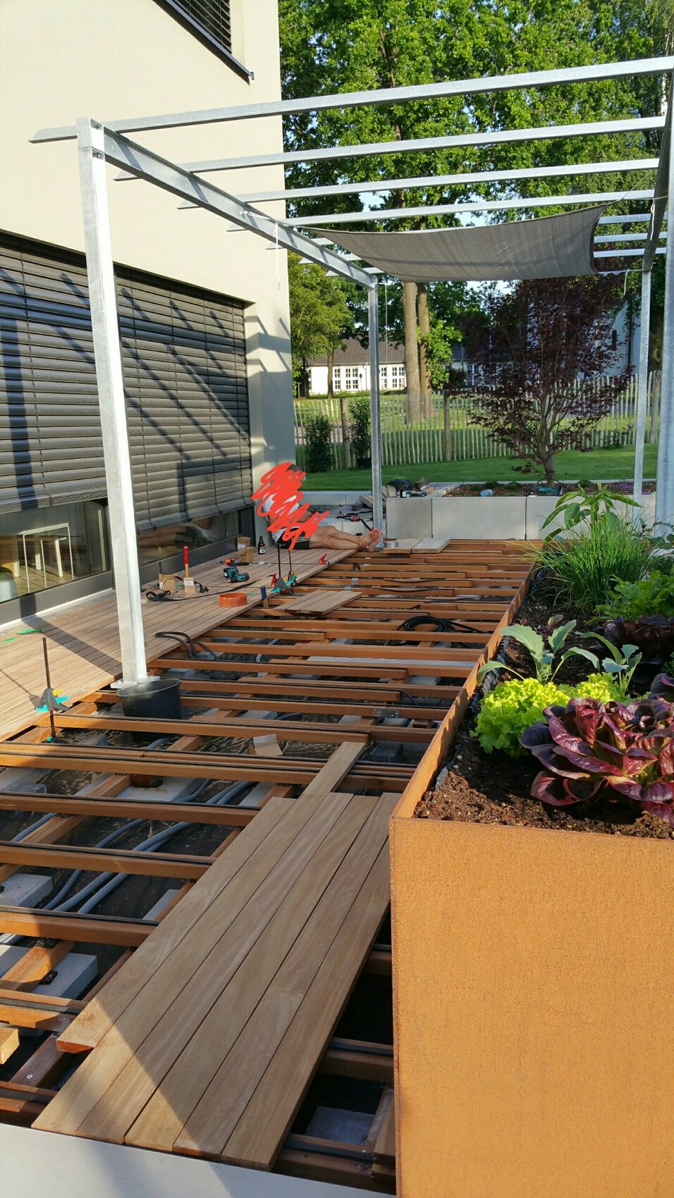 Wood terrace construction: metal frame, wooden decking, tools; sun sail, garden bed on the right.