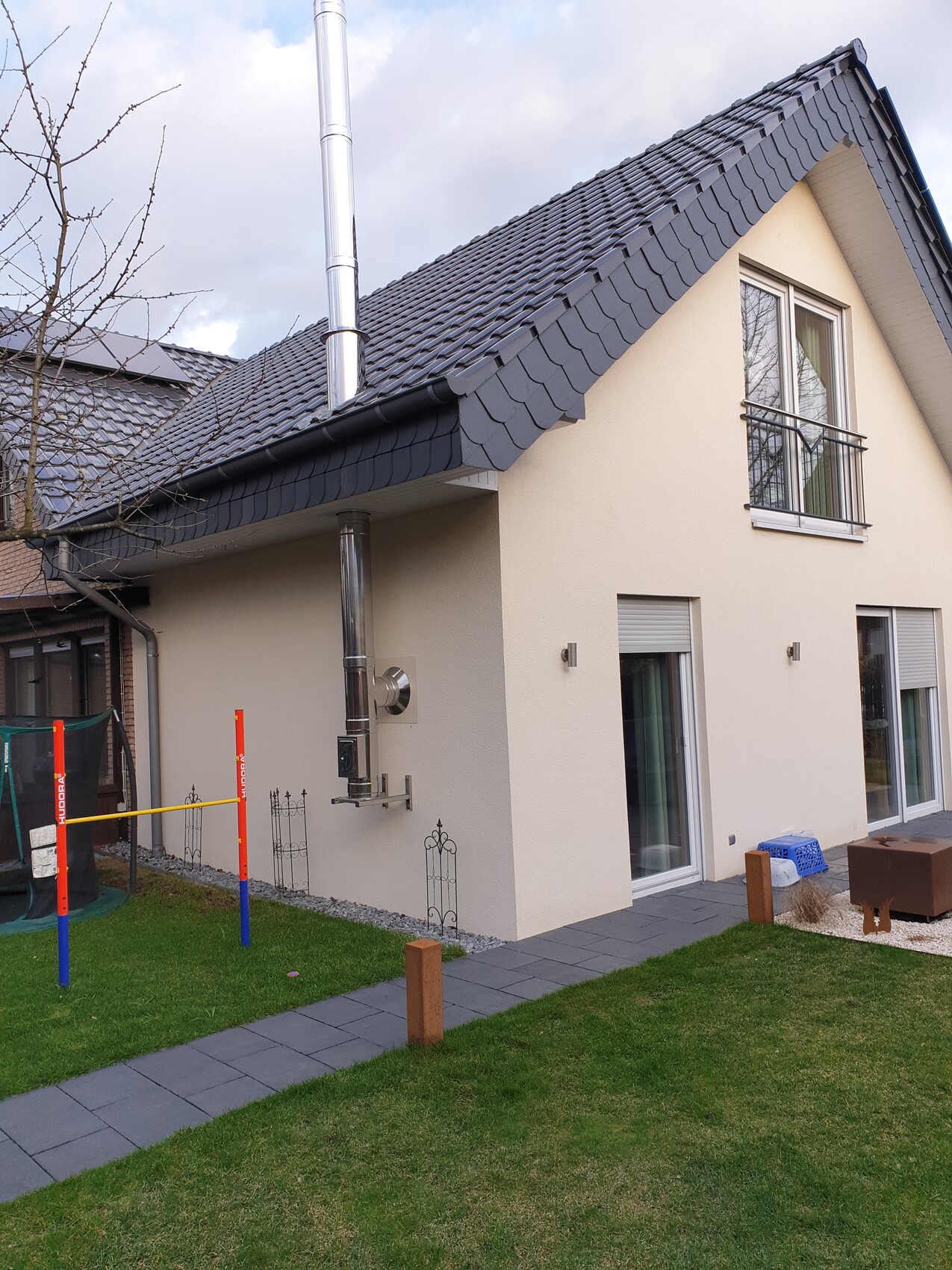 Modern beige detached house with dark roof, metal chimney, terrace, and trampoline in the garden.
