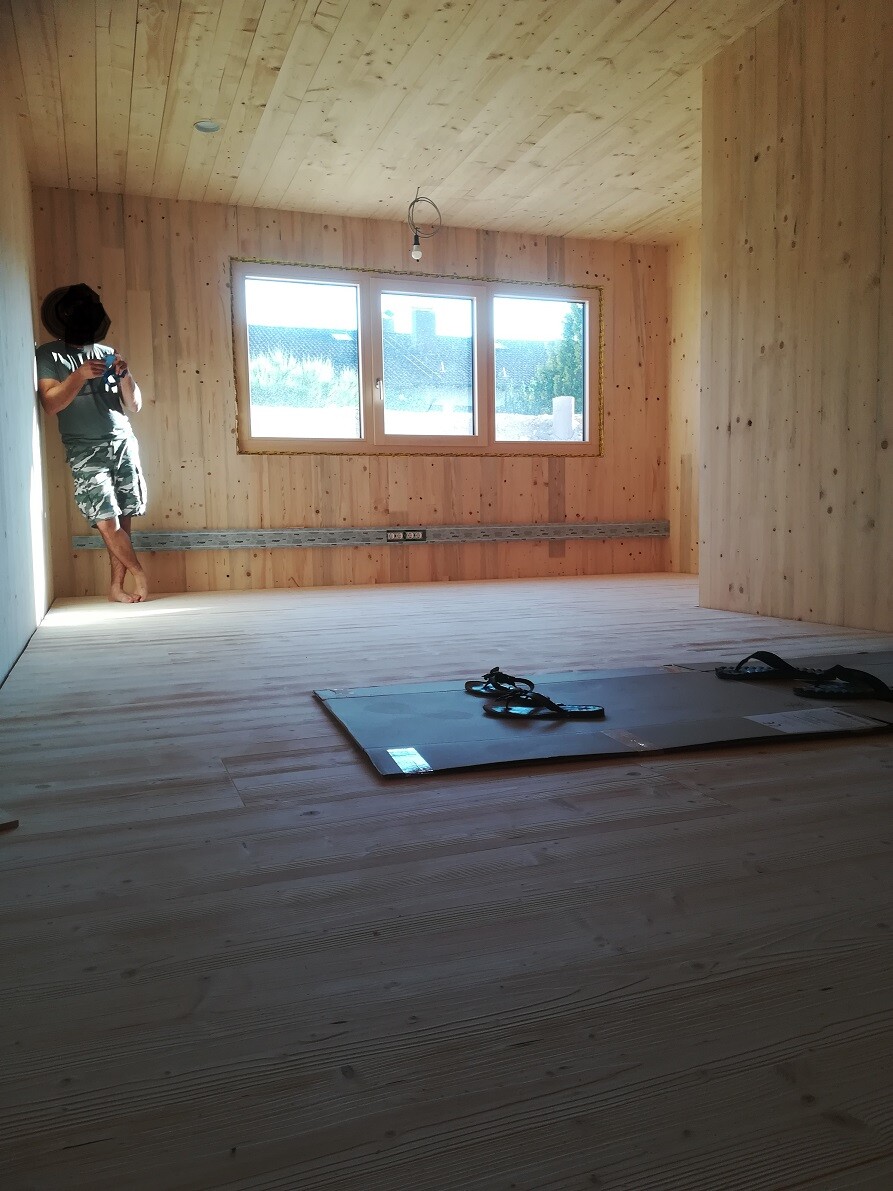 Unfinished wooden room under construction; man in shorts leaning against wall next to window, tools on the floor.