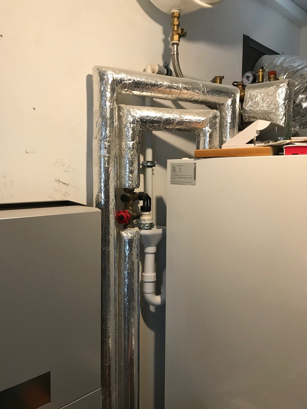 Heating room with insulated pipes, boiler and red shut-off valve next to white cabinet.