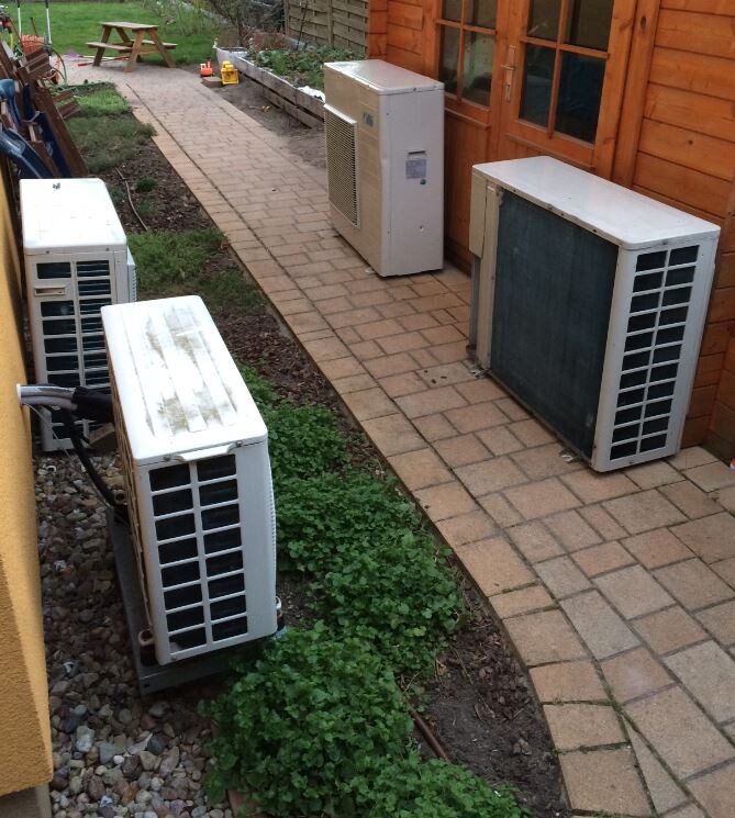 Several outdoor air conditioning units are standing on a paved path next to a wooden house.