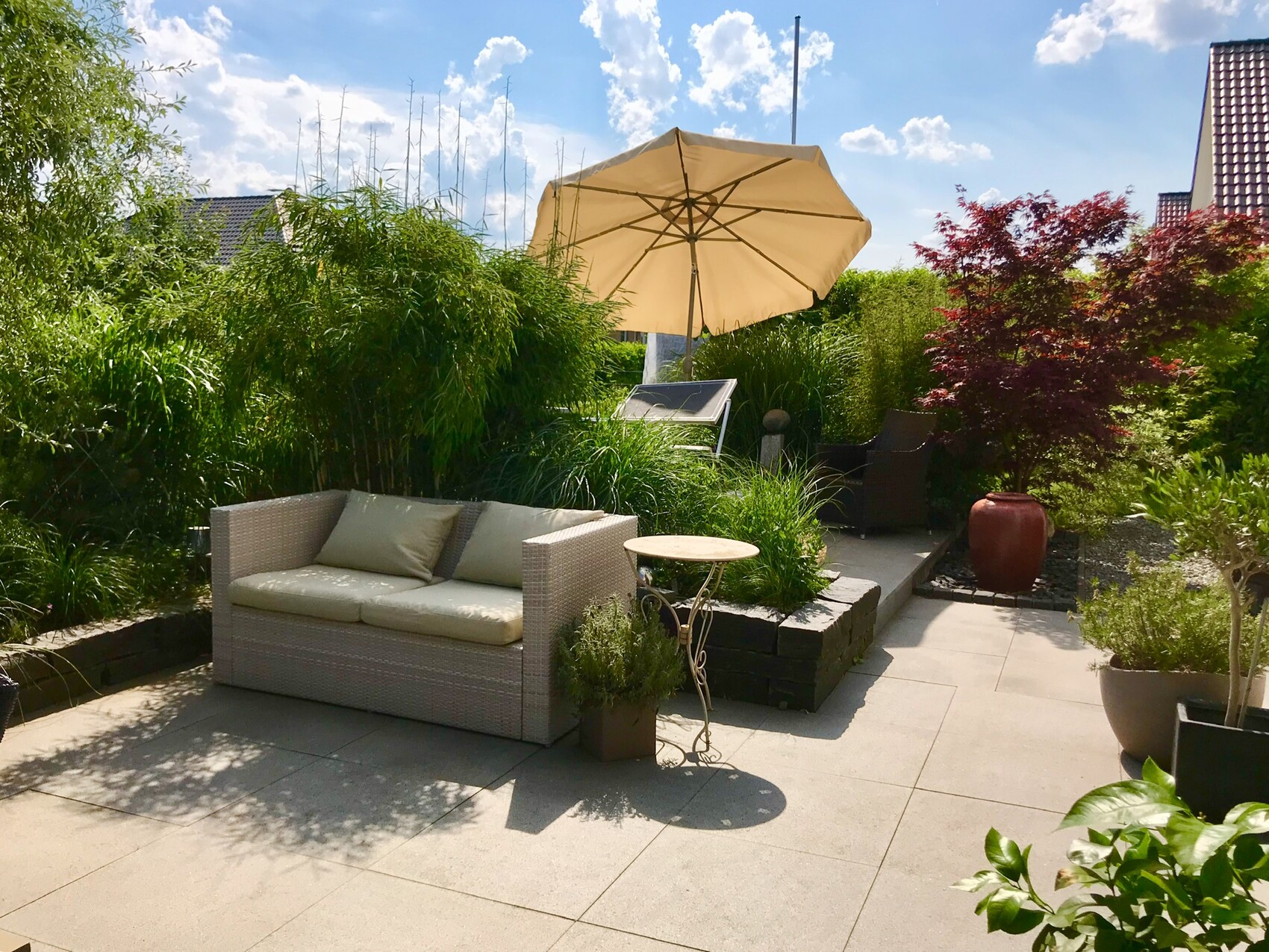 Garden terrace with light rattan sofa, sunshade, table, many plants and trees.