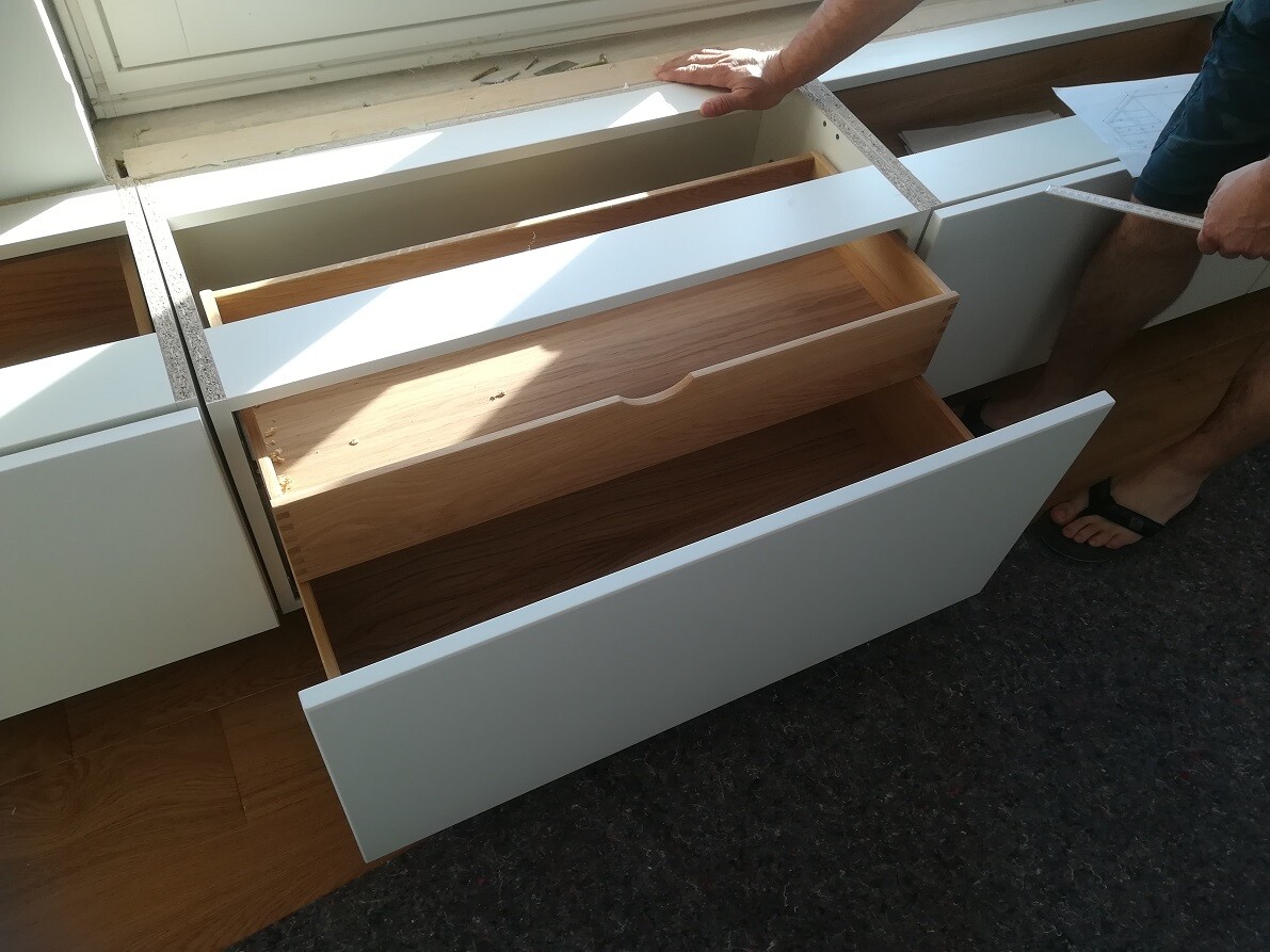 Person placing tape measure on white kitchen drawers; several drawers open, wooden interior visible.