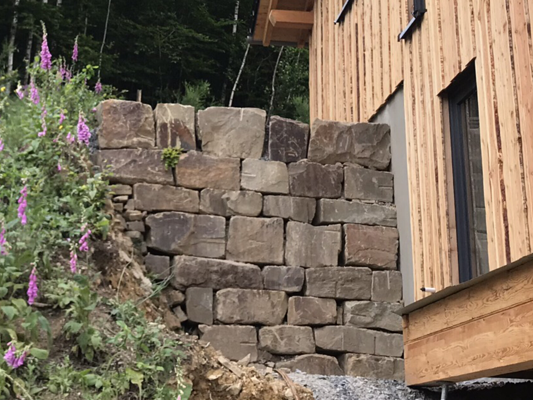 Large natural stone retaining wall next to a wooden house; left side plants with purple flowers.