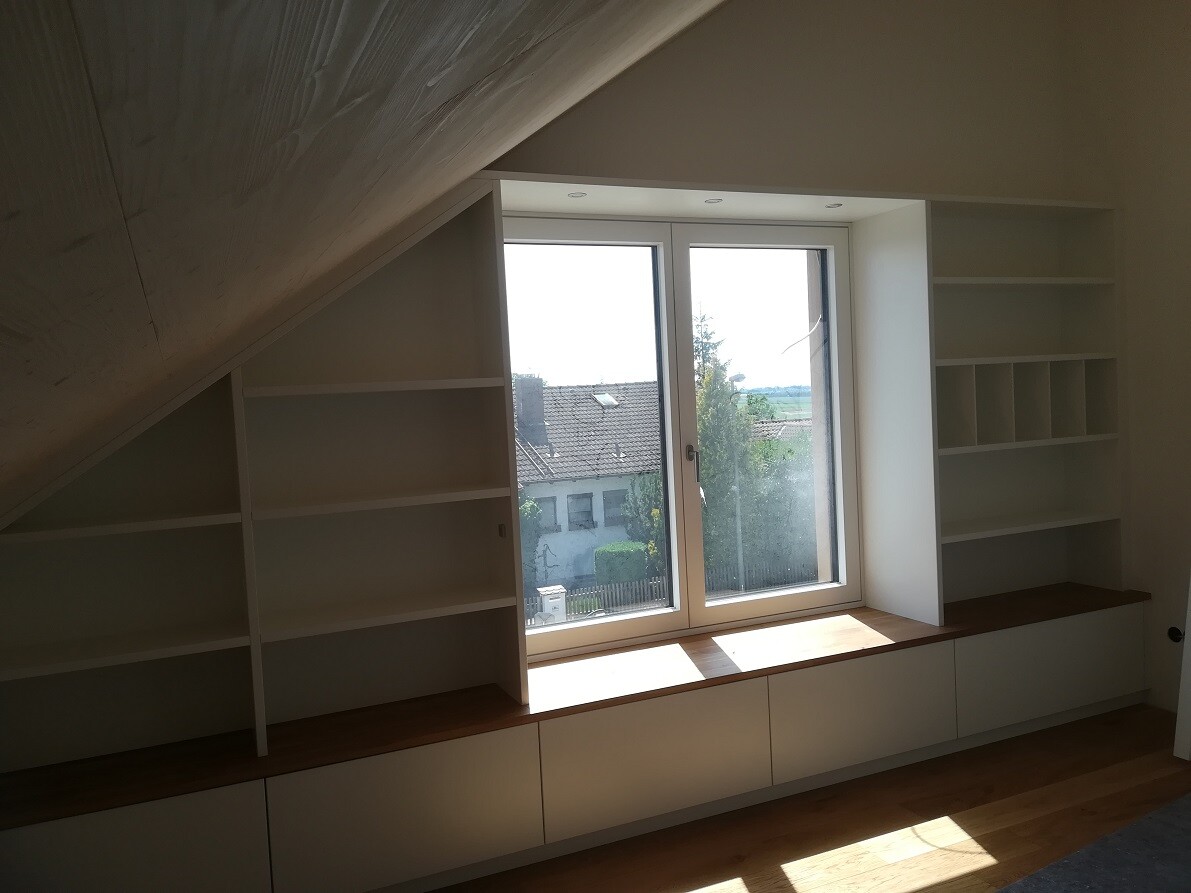 Bright room with sloped ceiling, white shelves surrounding a large window with a windowsill and base cabinets.