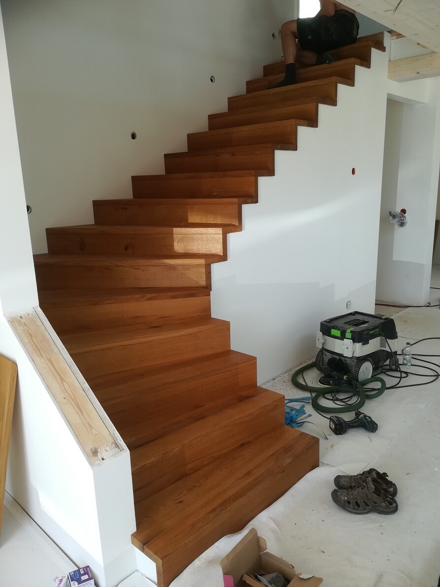 Modern wooden staircase indoors, construction work; tools, shoes, and materials on the floor; person at the top.
