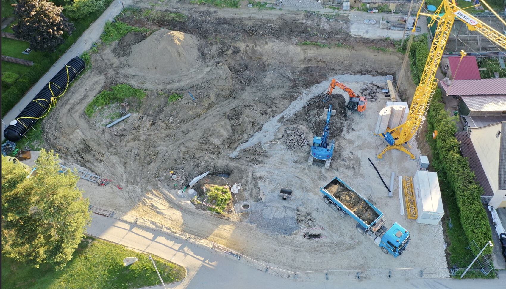 Luftaufnahme einer Baustelle: großer Erdaushub, gelber Turmkran, Bagger und LKW.