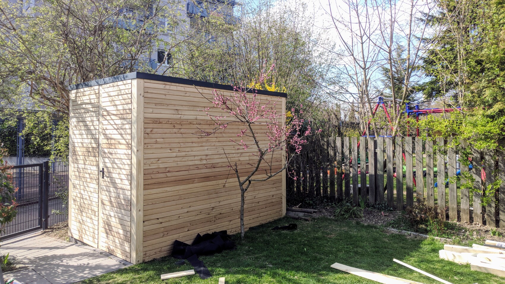 Garden with a light-colored shed on the left, blooming tree with pink flowers, fence on the right, playground behind