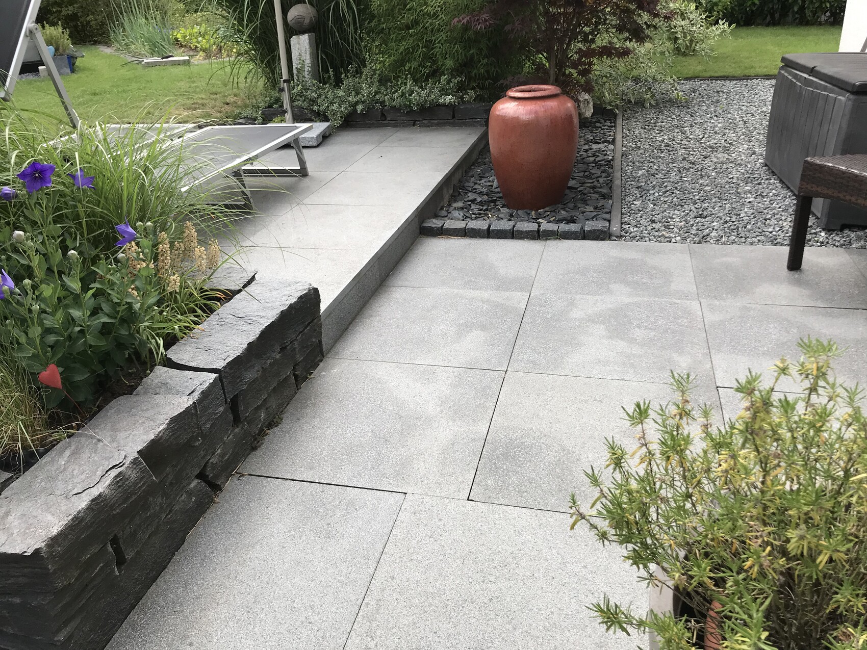 Gepflasterte Terrasse mit grauen Platten, roter Keramiktopf auf Kies, links Staudenbeet.