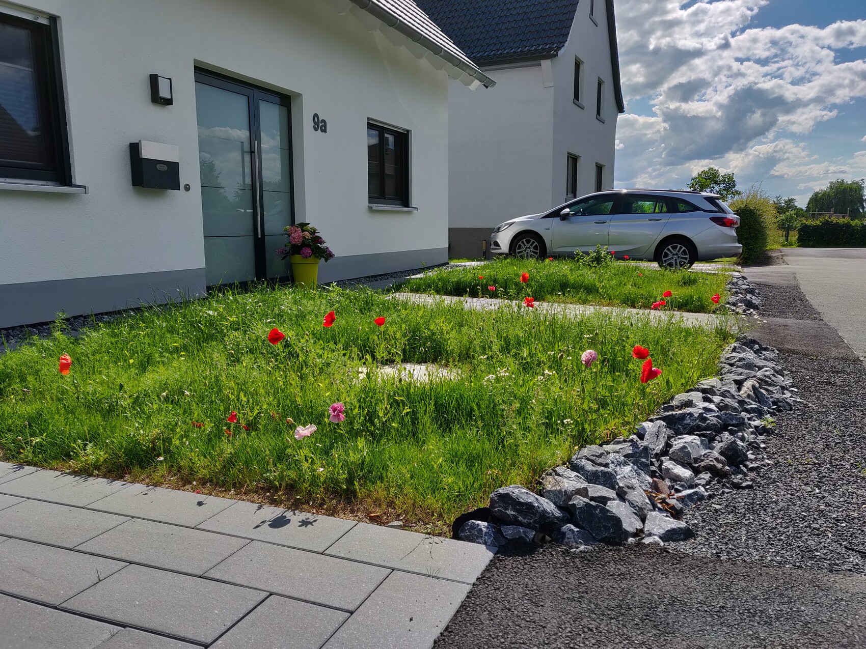 White house with gray door, yellow flowerpot, red poppies in the garden, and silver car.