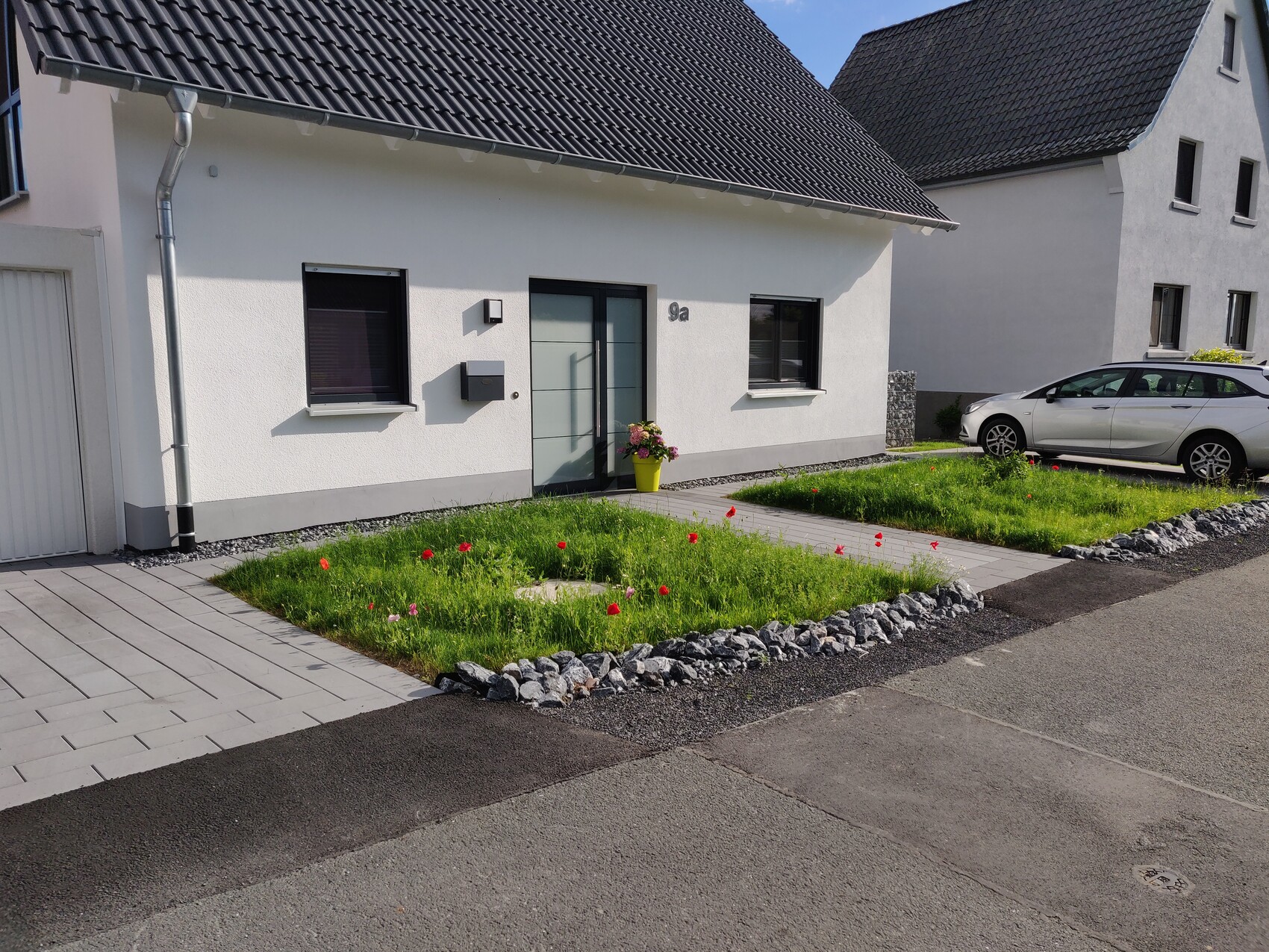 White detached house with dark roof, garden with tulips and yellow flowerpot, silver passenger car.