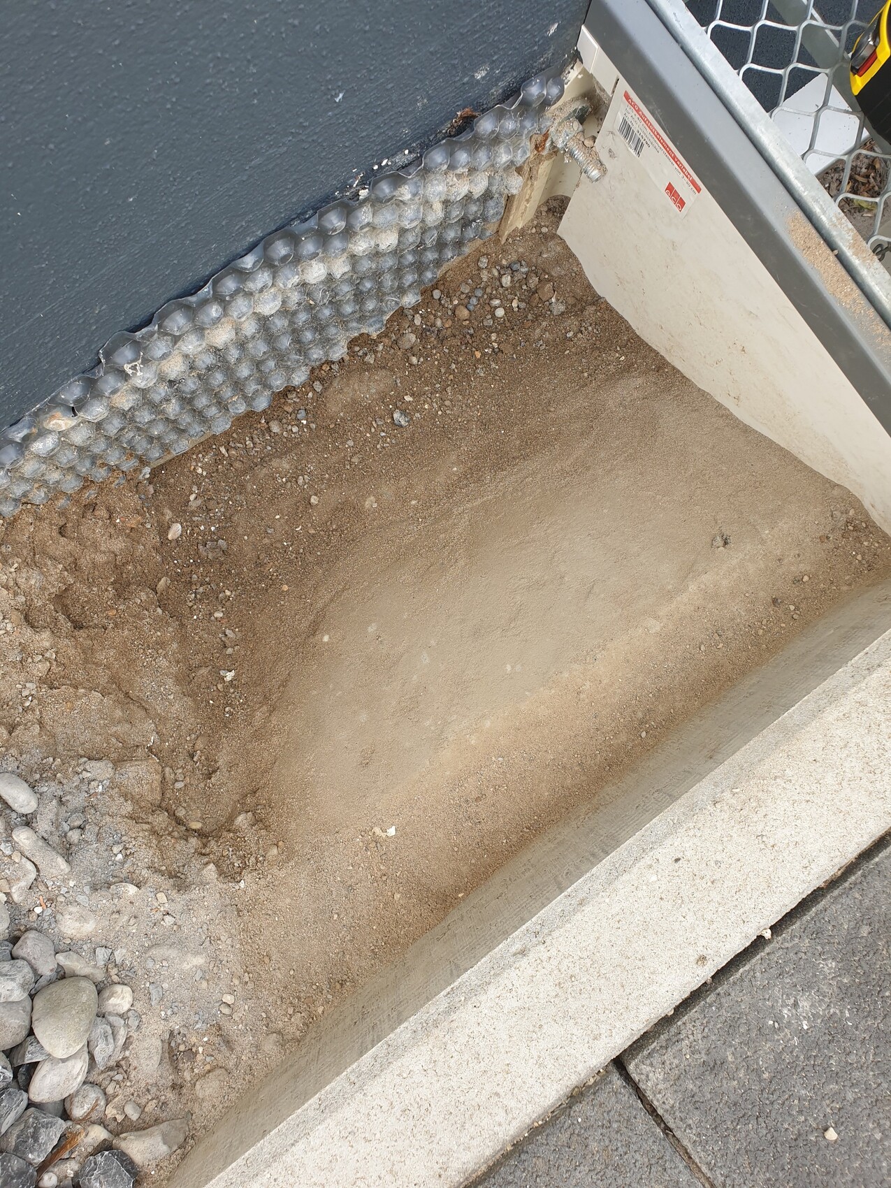 Bodenaushub neben Gebäudefundament; Sand, Kies in einer Baugrube; Betonrand sichtbar.