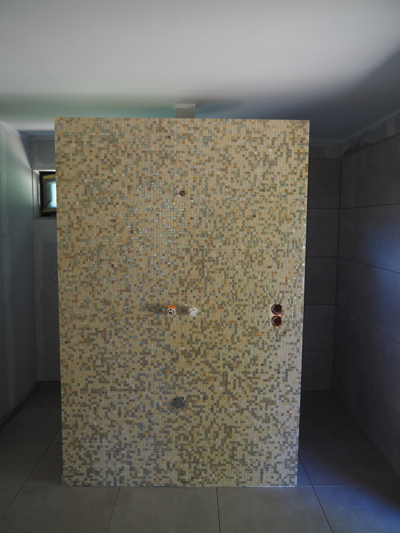 Beige mosaic tile wall made of small tiles in the bathroom, water connections visible.