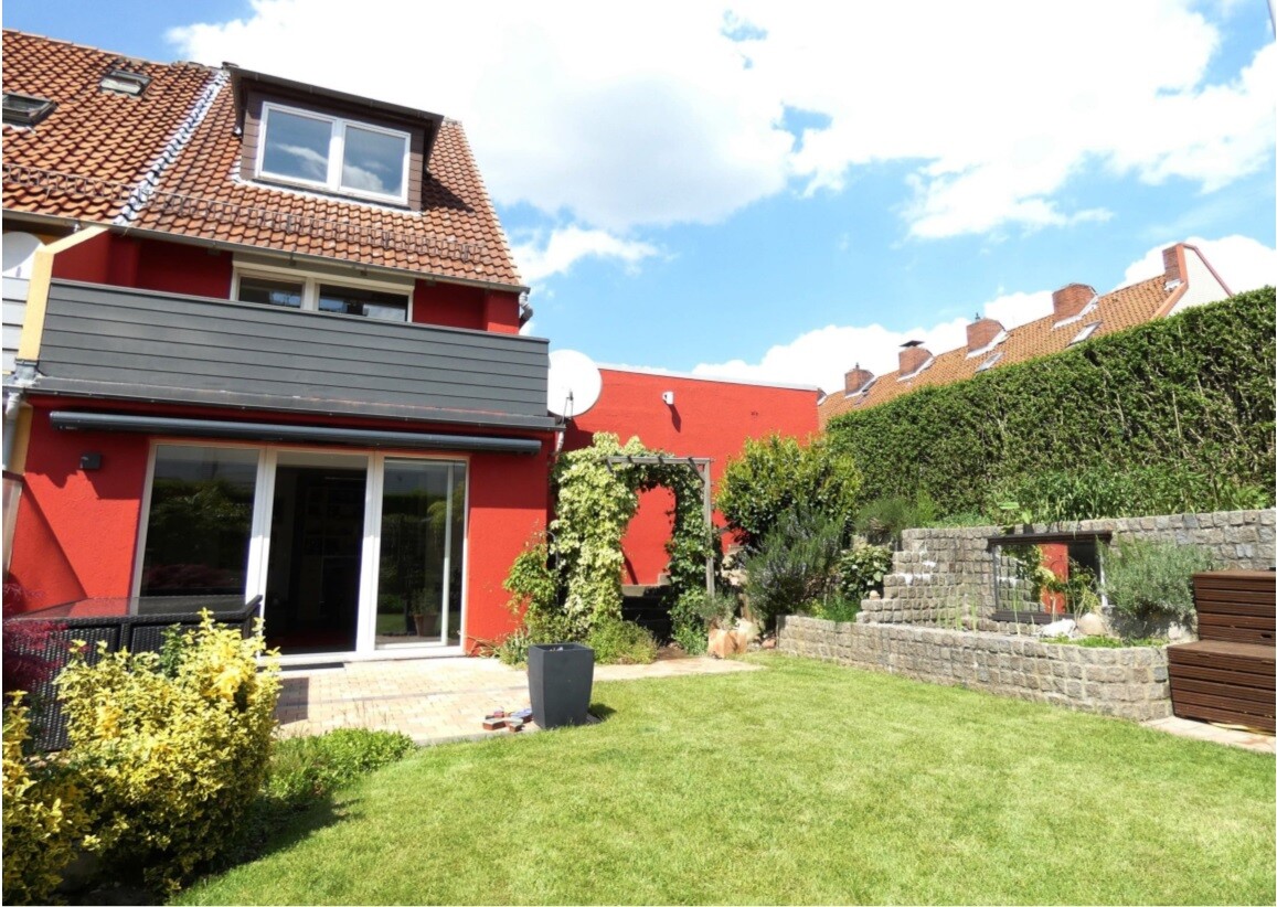 Garden behind red house: terrace with glass sliding door, balcony, green hedge and stone wall.