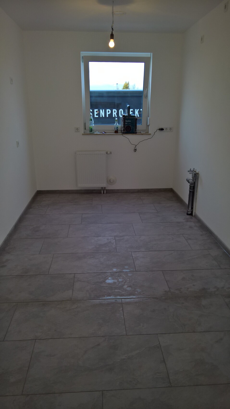 Bright, empty room with tiled floor, hanging light bulb, window, radiator, and pipework.