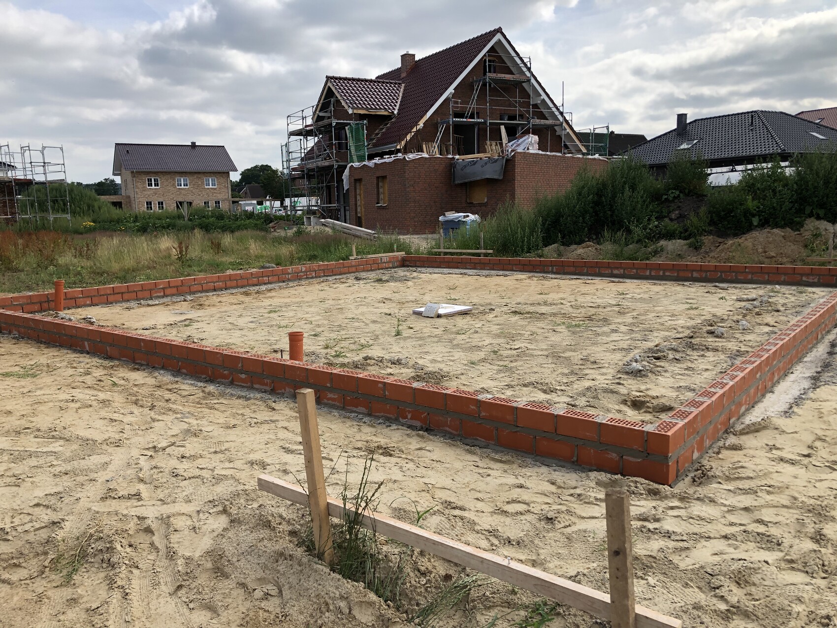 Building plot with rectangular brick wall on sand; house with scaffolding in the background.