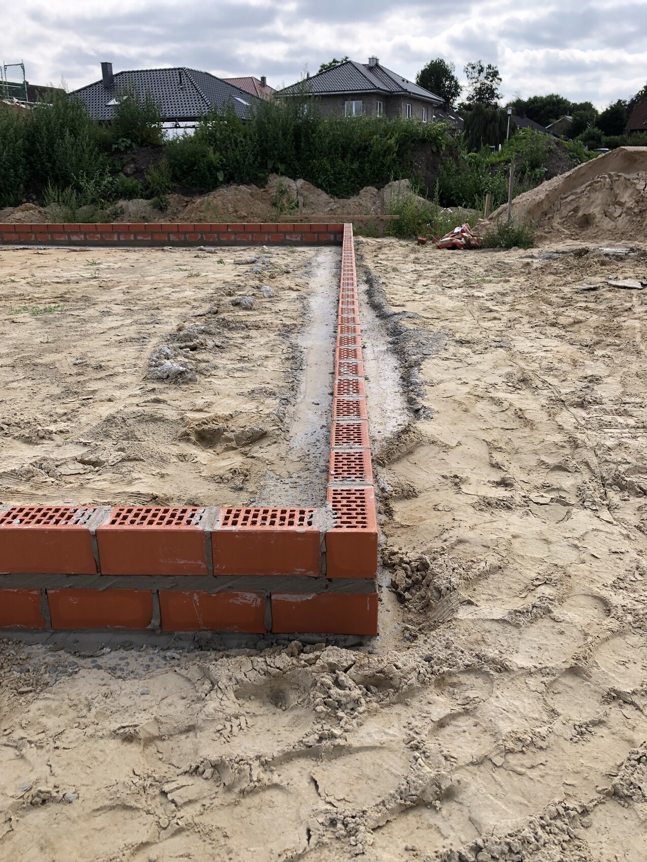 Construction area: long row of red perforated bricks in a trench, sandy soil, houses in the background.