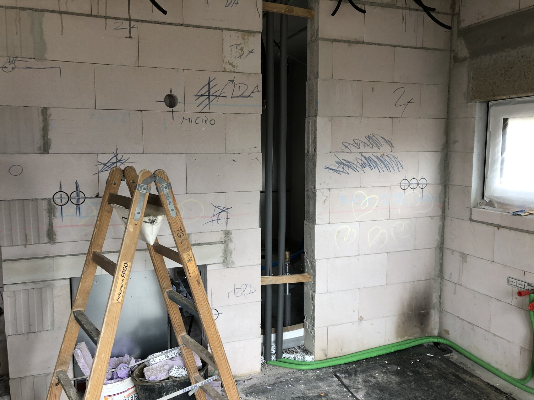 Construction site: unfinished block wall, wooden ladder, green hose on the floor, graffiti, window on the right.