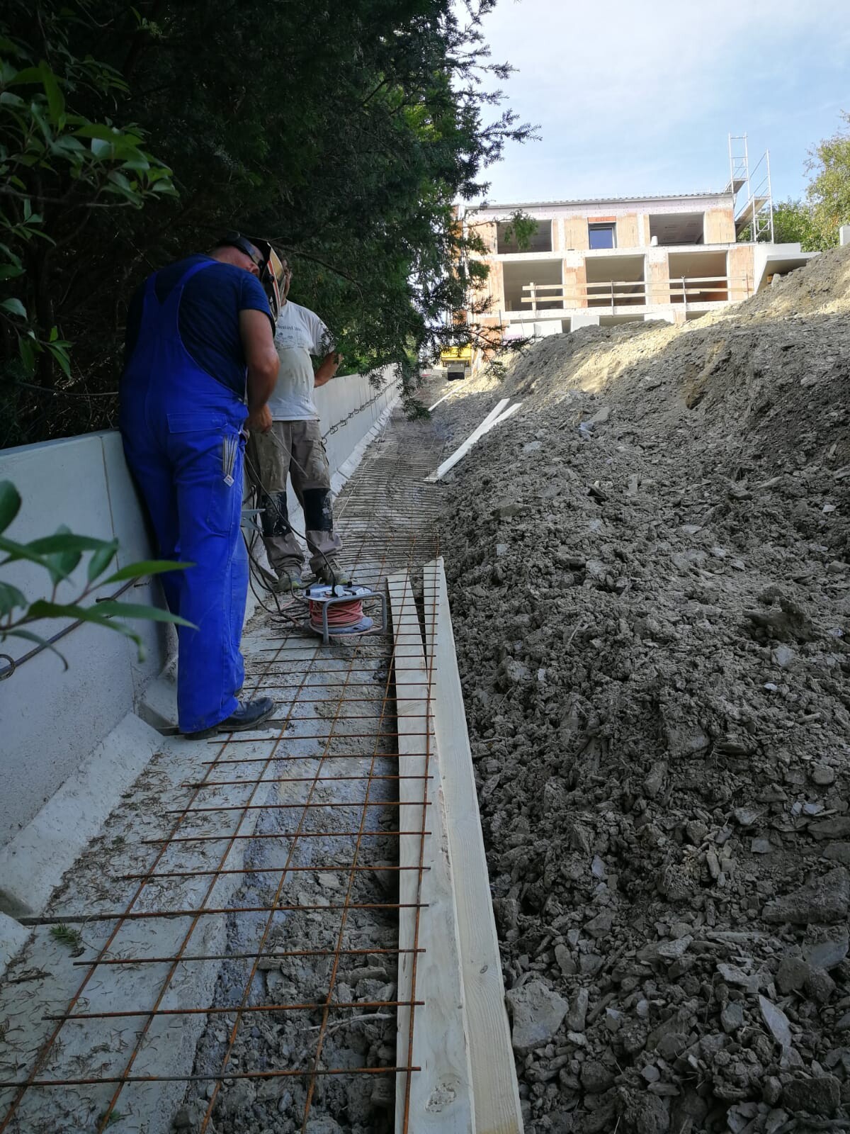 Two construction workers installing reinforcement on concrete footing formwork; earth and gravel pile to the right, shell structure.