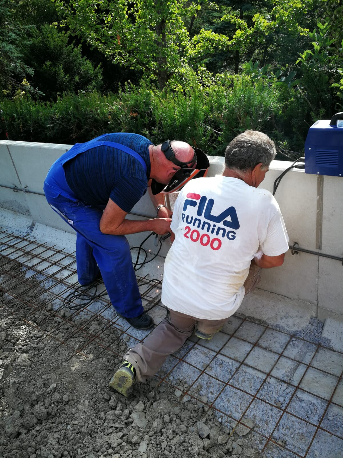 Two construction workers placing steel reinforcement in outdoor concrete formwork, one wearing a face shield.