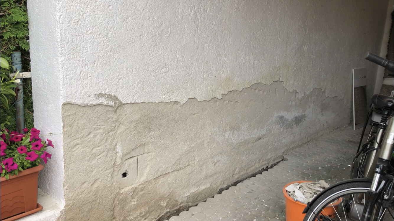 Exterior wall with peeling plaster, grey substrate at the bottom; flower pot on the left, bicycle on the right.