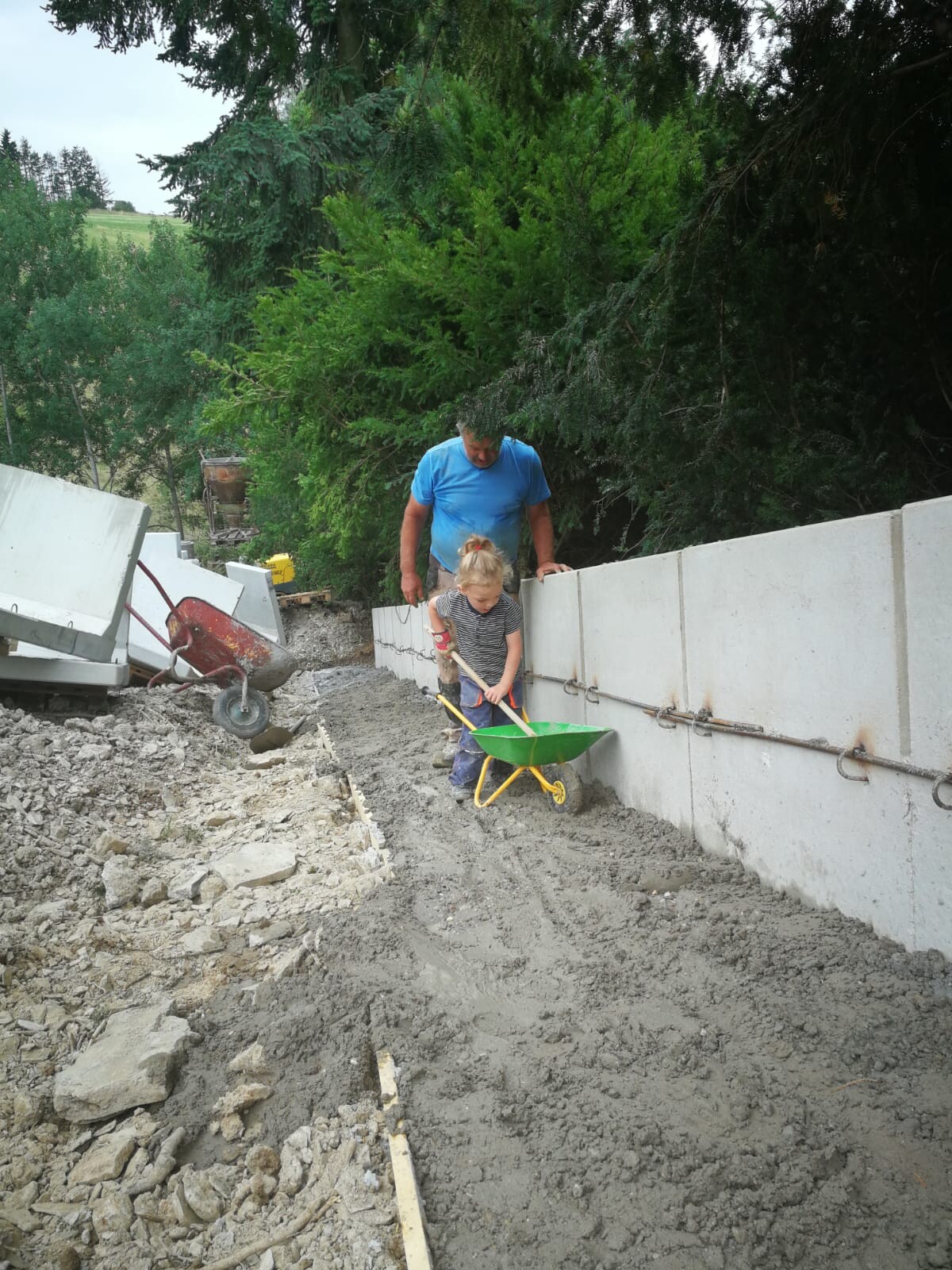 A man in a blue shirt helping a child dig with a green wheelbarrow next to a concrete wall.