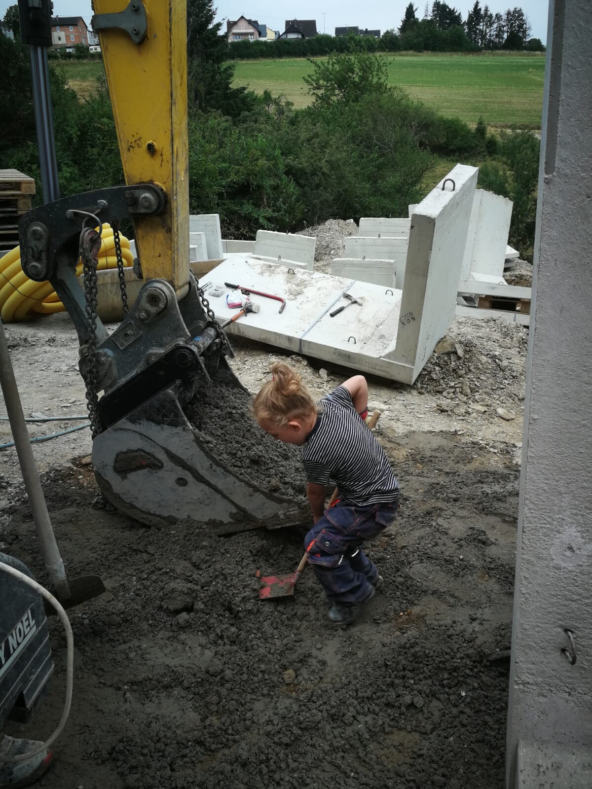 Small child digging with a shovel in the mud next to a yellow excavator on a construction site.