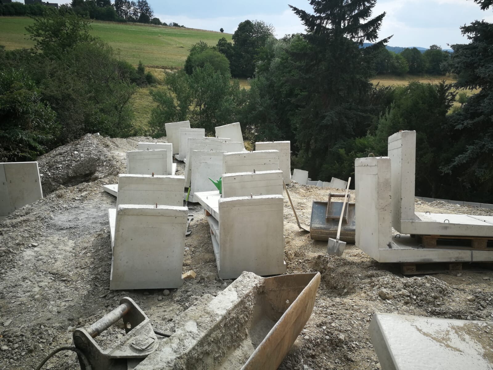 Construction site with concrete foundations and supports, debris, construction tools; background: fields and trees
